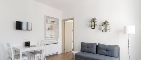Una sala de estar moderna con un sofá gris, una mesa de comedor blanca con sillas, un televisor de pantalla plana y una estantería.