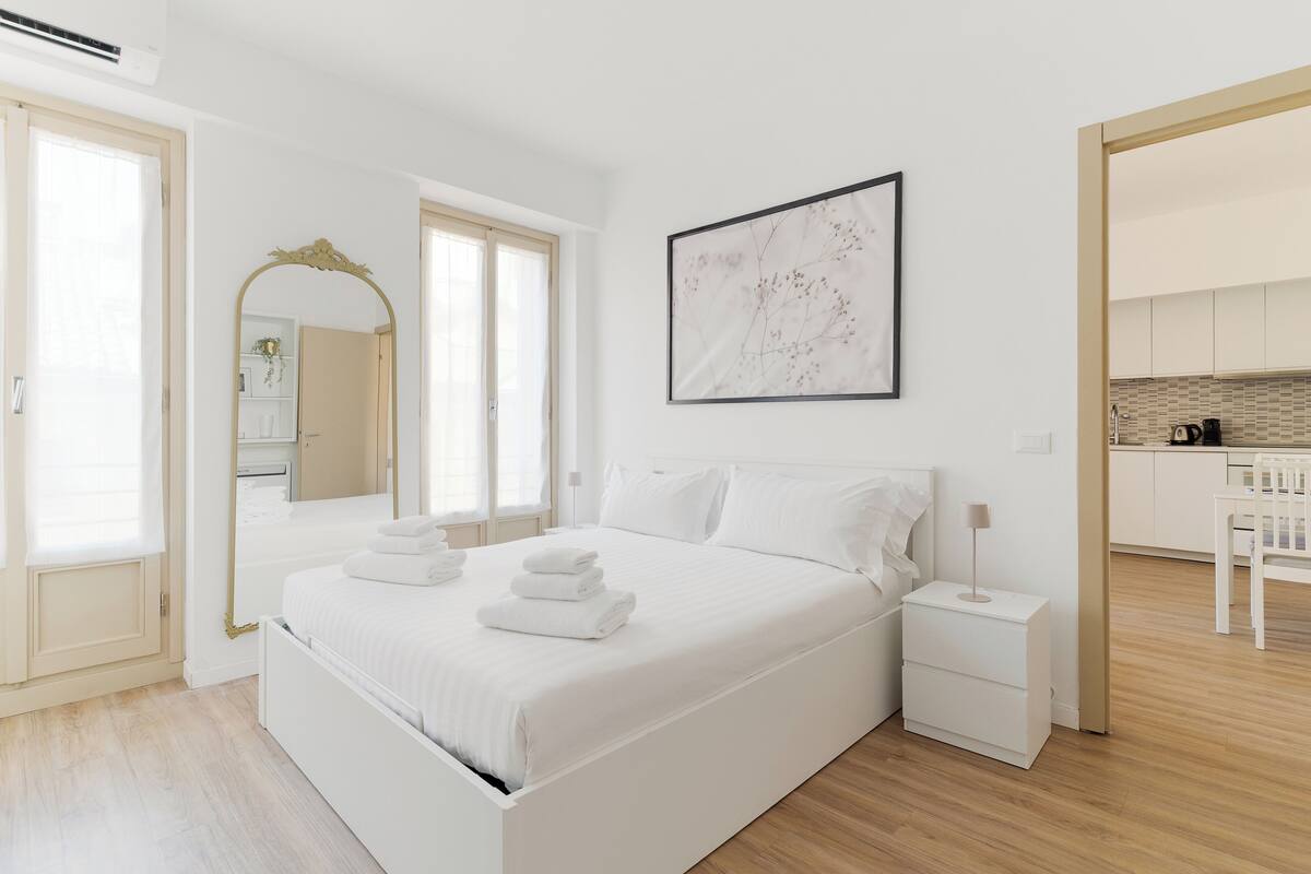 A modern bedroom with a large bed, bedside tables, a mirror, and a view into the kitchen.