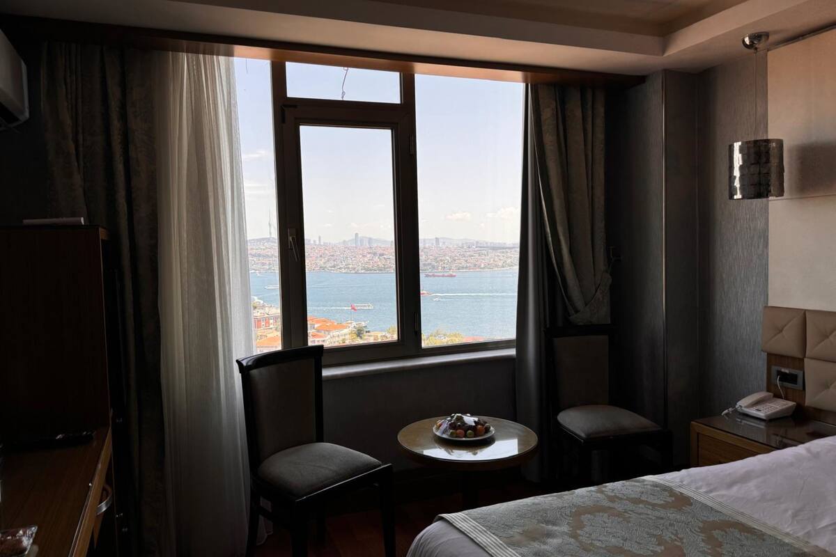 Habitación de hotel con un ventanal amplio que ofrece vistas a la ciudad, una cama, una mesita con una fuente de frutas y una silla.