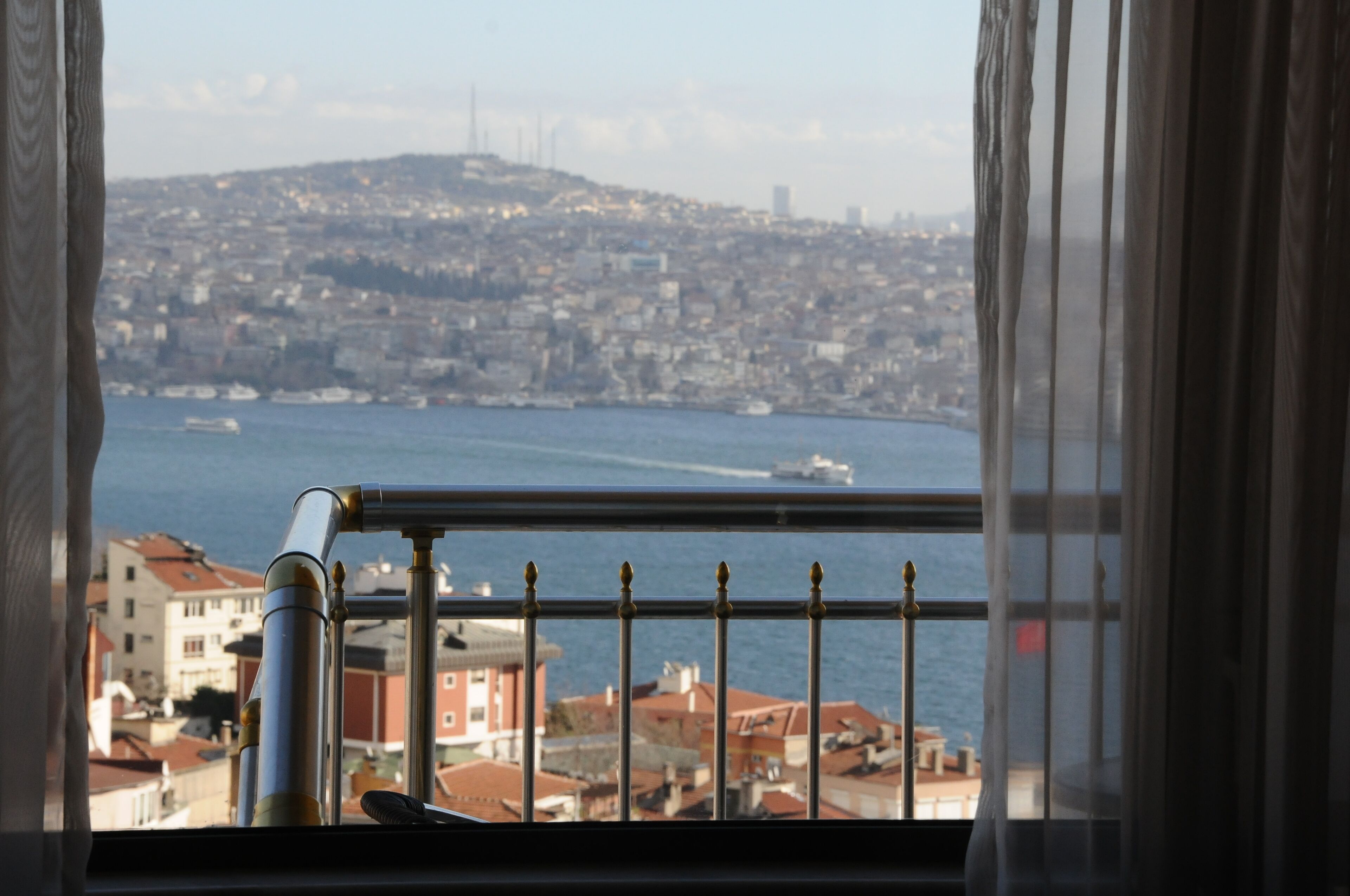 A view from a window overlooking a cityscape with a body of water, a hill in the distance, and a railing in the foreground.