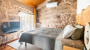Un dormitorio con pared de piedra, techo de madera, cama con ropa de cama estampada, una mesita de noche y un televisor sobre un mueble.