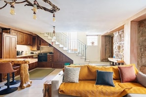 A modern kitchen with wooden cabinets, a dining area with a wooden table and chairs, a staircase leading to another level, and a cozy living area with a sofa and cushions.