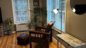 Uma sala de estar com piso de madeira, uma poltrona de couro marrom, uma mesa pequena e um abajur.