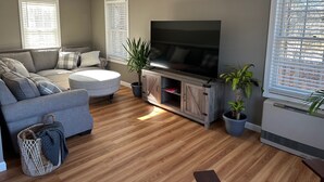 Uma sala de estar com um sofá cinzento, uma mesa de centro redonda, uma televisão de tela plana sobre um suporte de madeira e plantas em vasos.
