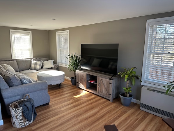 A living room with a grey sofa, a round coffee table, a flat-screen TV on a wooden stand, and potted plants. - Beautiful Private Home in the Heart of Wells, ME! Only 1.6mi from the beach! (Wells)