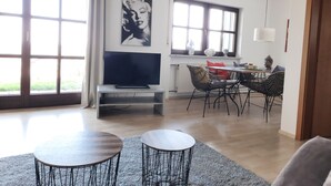 Un salon moderne doté d’un téléviseur à écran plat, d’un coin repas avec une table et des chaises, et de grandes fenêtres avec des rideaux.