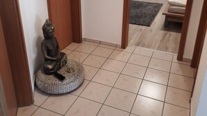 Un couloir avec un sol carrelé, une porte en bois, une statue et, au fond, un lit recouvert d’une couvre-lit à motifs floraux.