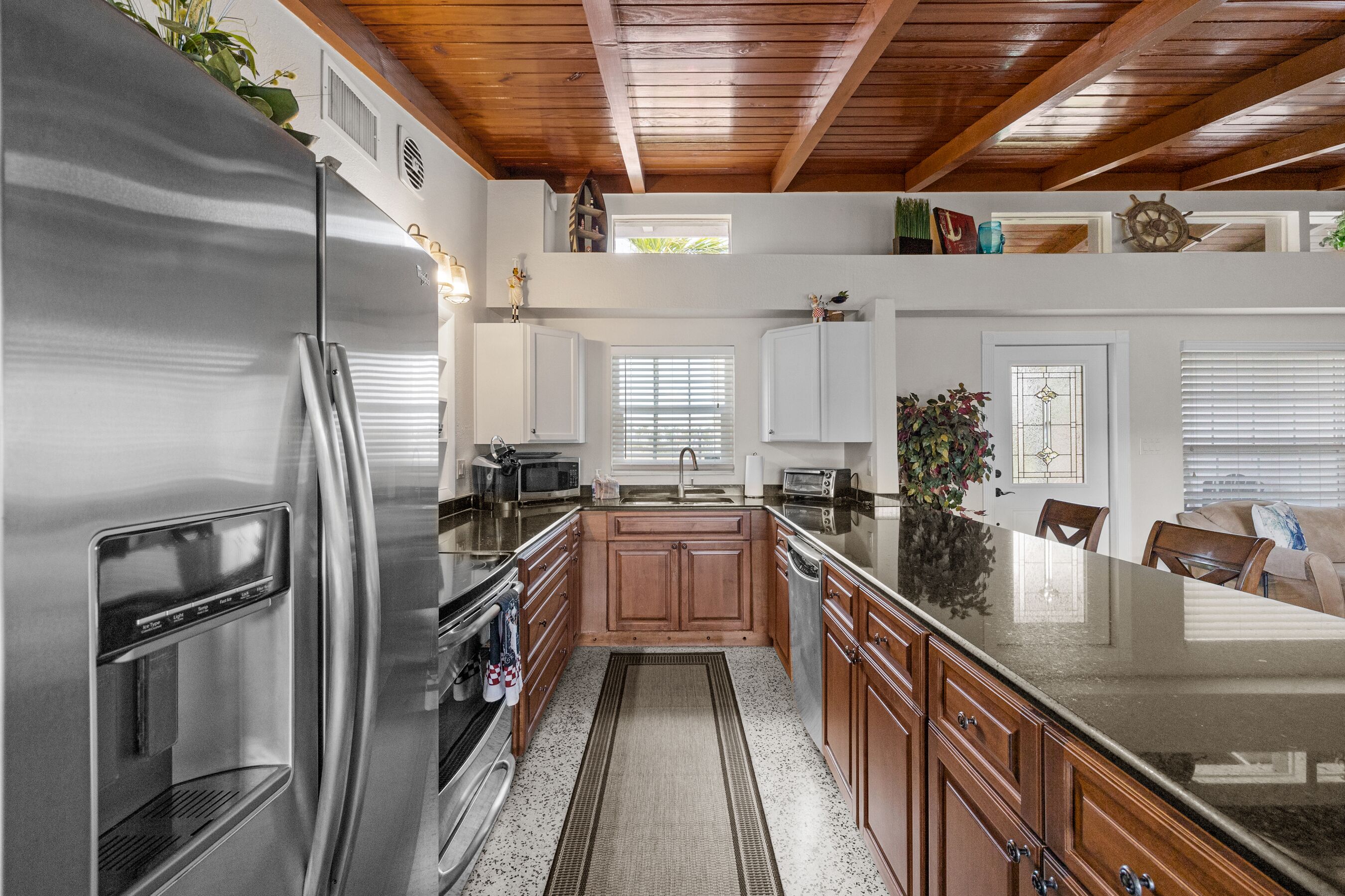 A kitchen with a stainless steel refrigerator, wooden cabinets, a granite countertop, and a window with blinds.