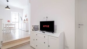 Ein modernes Wohnzimmer mit einem weißen Schrank, einem Flachbildfernseher, der 'NETFLIX' anzeigt, und einem Essbereich mit weißem Tisch und Stühlen.