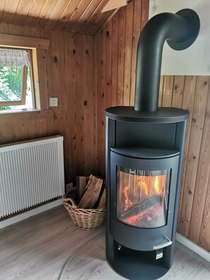 Ein moderner Holzofen mit Glastür, neben einem Fenster und einer Heizung in einem raumausgestatteten Holzzimmer.