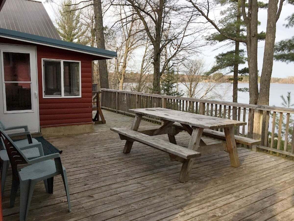 Une terrasse en bois avec des tables et des chaises pour pique-niquer, un petit chalet rouge et une vue sur un lac.