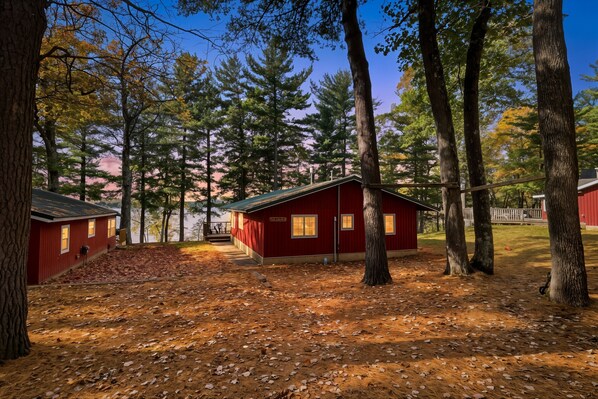 A cabin with a red exterior, surrounded by trees and a dirt ground.