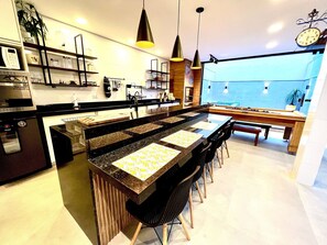 Una cocina moderna con una isla central, taburetes de bar, iluminación colgante y vista a una piscina por la ventana.