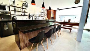 A modern kitchen with a central island, black chairs, pendant lighting, and a view of the city through large windows. - Casas Marimar Ilheus (Caraguatatuba)