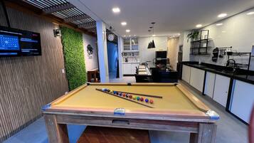 Une table de billard moderne pour l’intérieur, avec un jeu de billard (queue et billes), un téléviseur affichant l’interface Netflix, un mur vert orné d’une horloge, et un coin cuisine équipé d’appareils électroménagers et d’ustensiles.