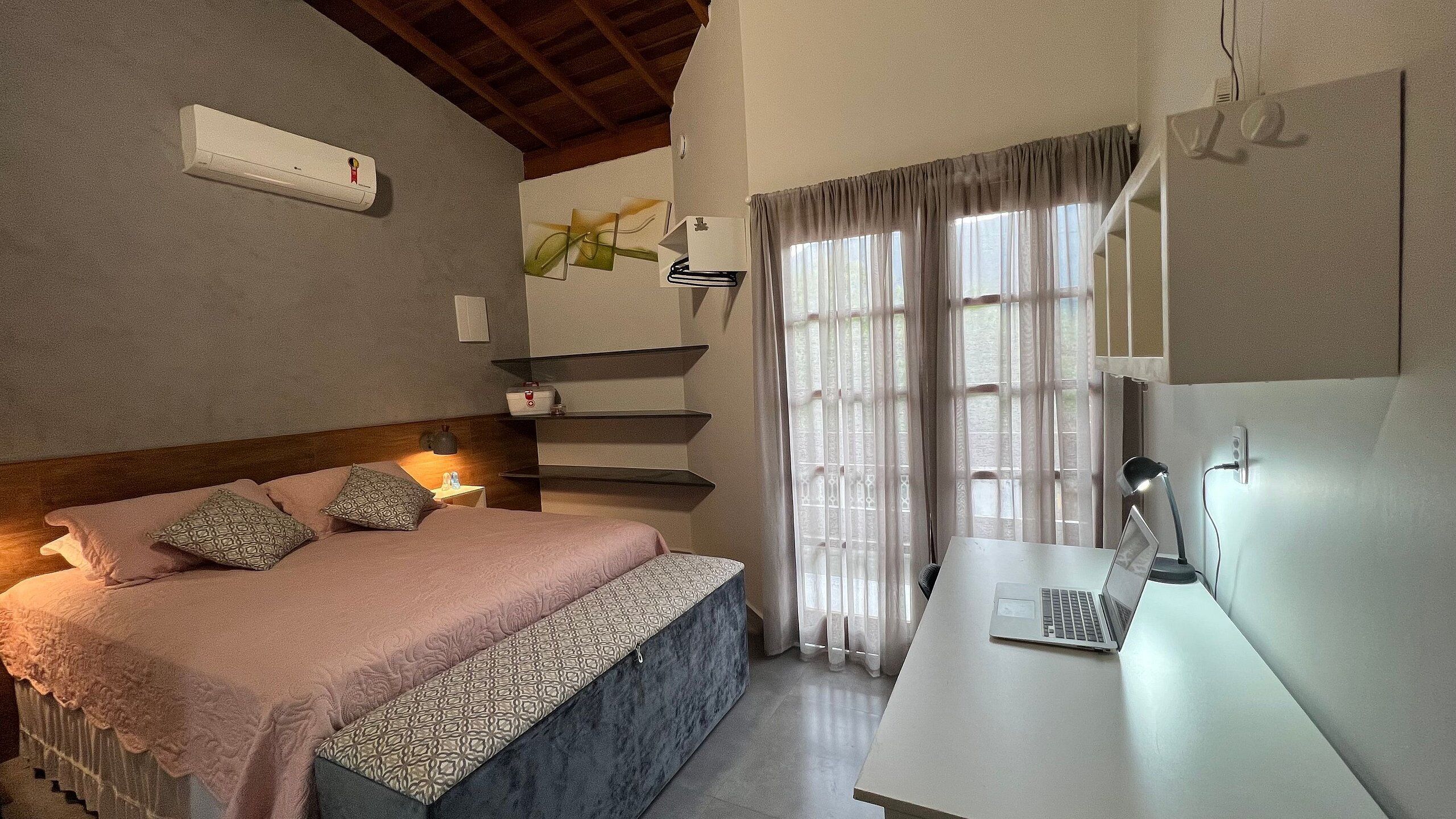 A bedroom with a bed, bedside tables, a desk with a laptop, a wall-mounted air conditioner, and a window with curtains.