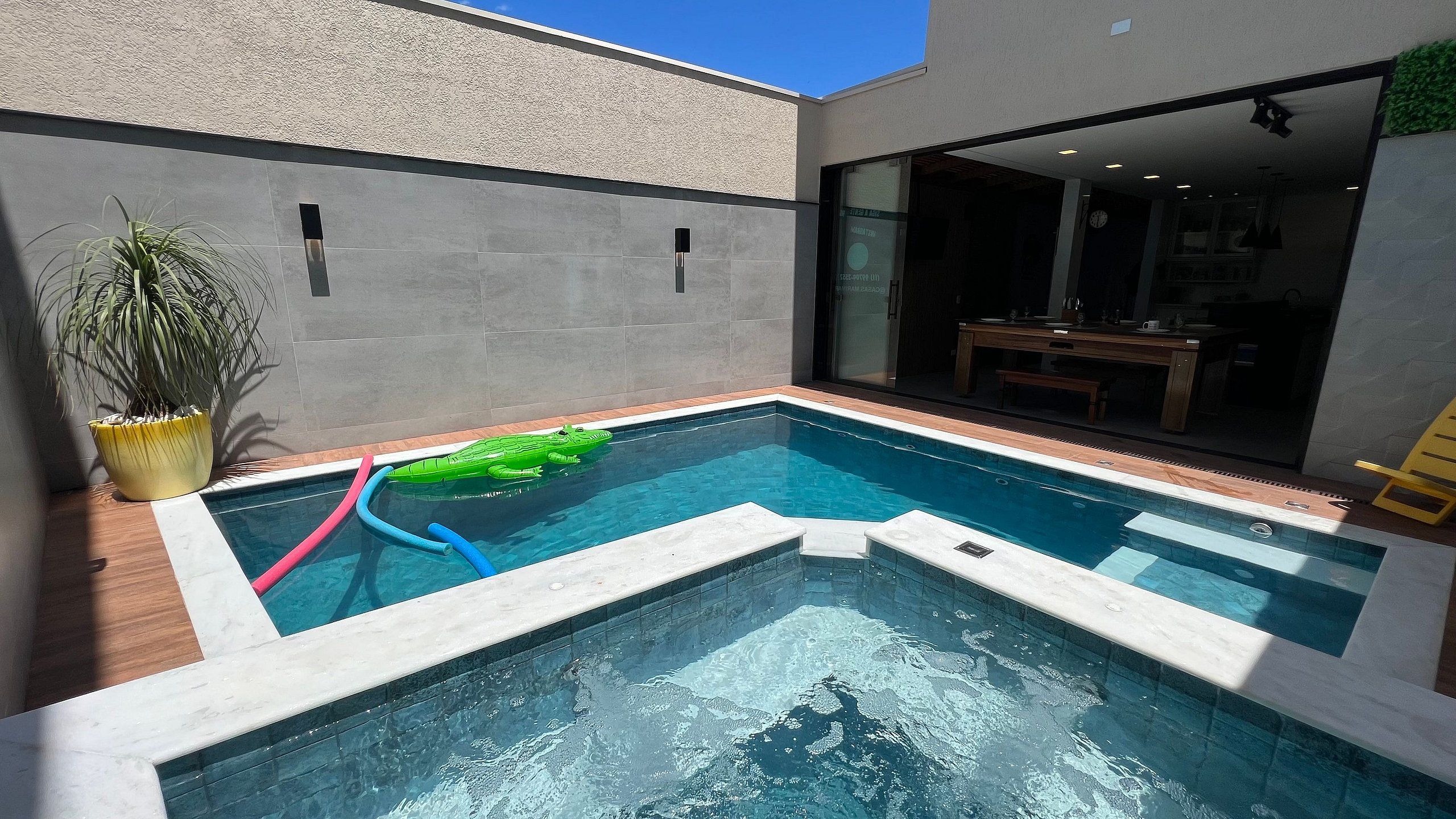 A swimming pool with a small inflatable toy, a potted plant, and a view of an indoor area with a table and chairs.