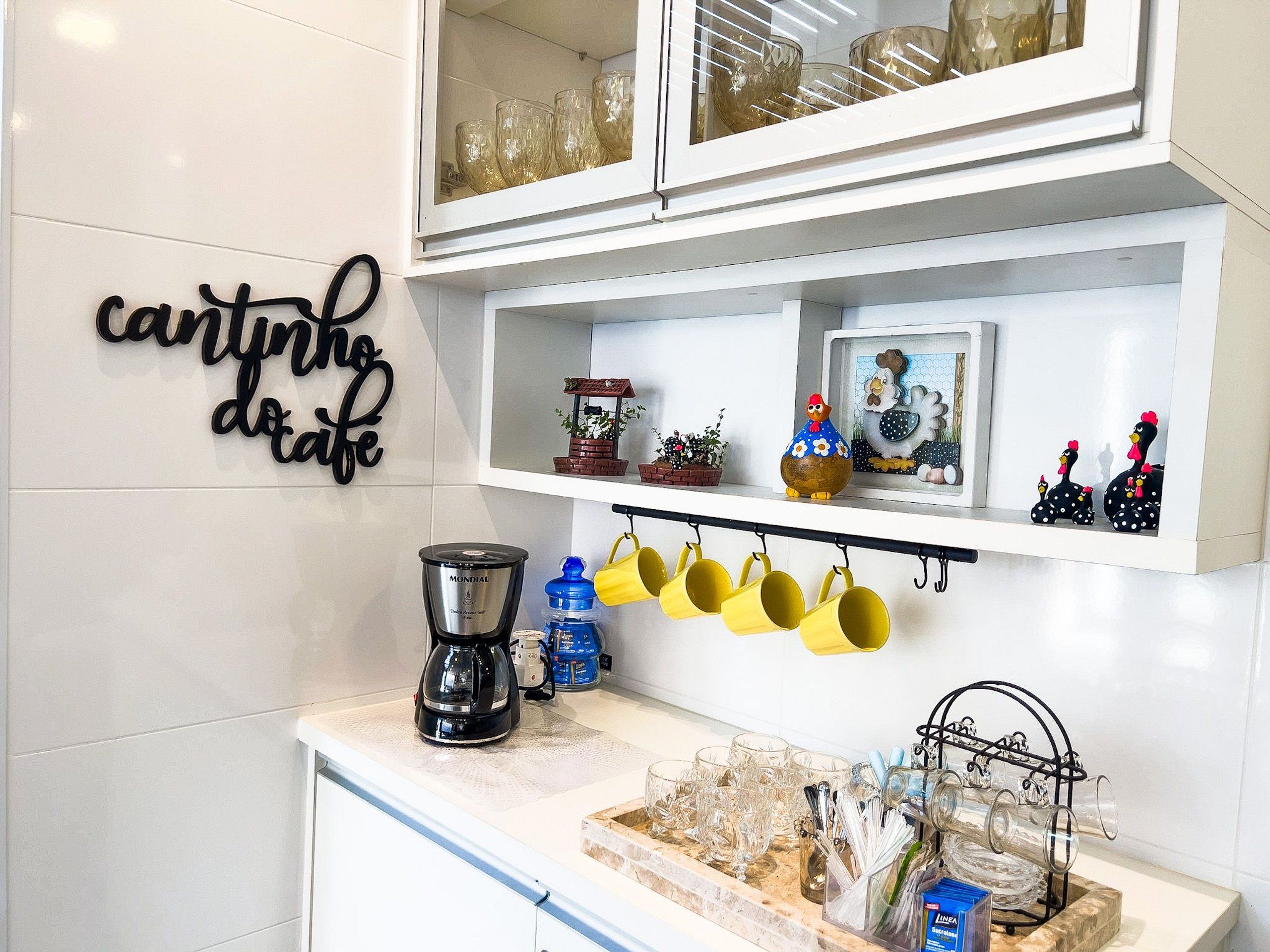 Una cocina con armarios blancos, cafetera, tazas amarillas y elementos decorativos.