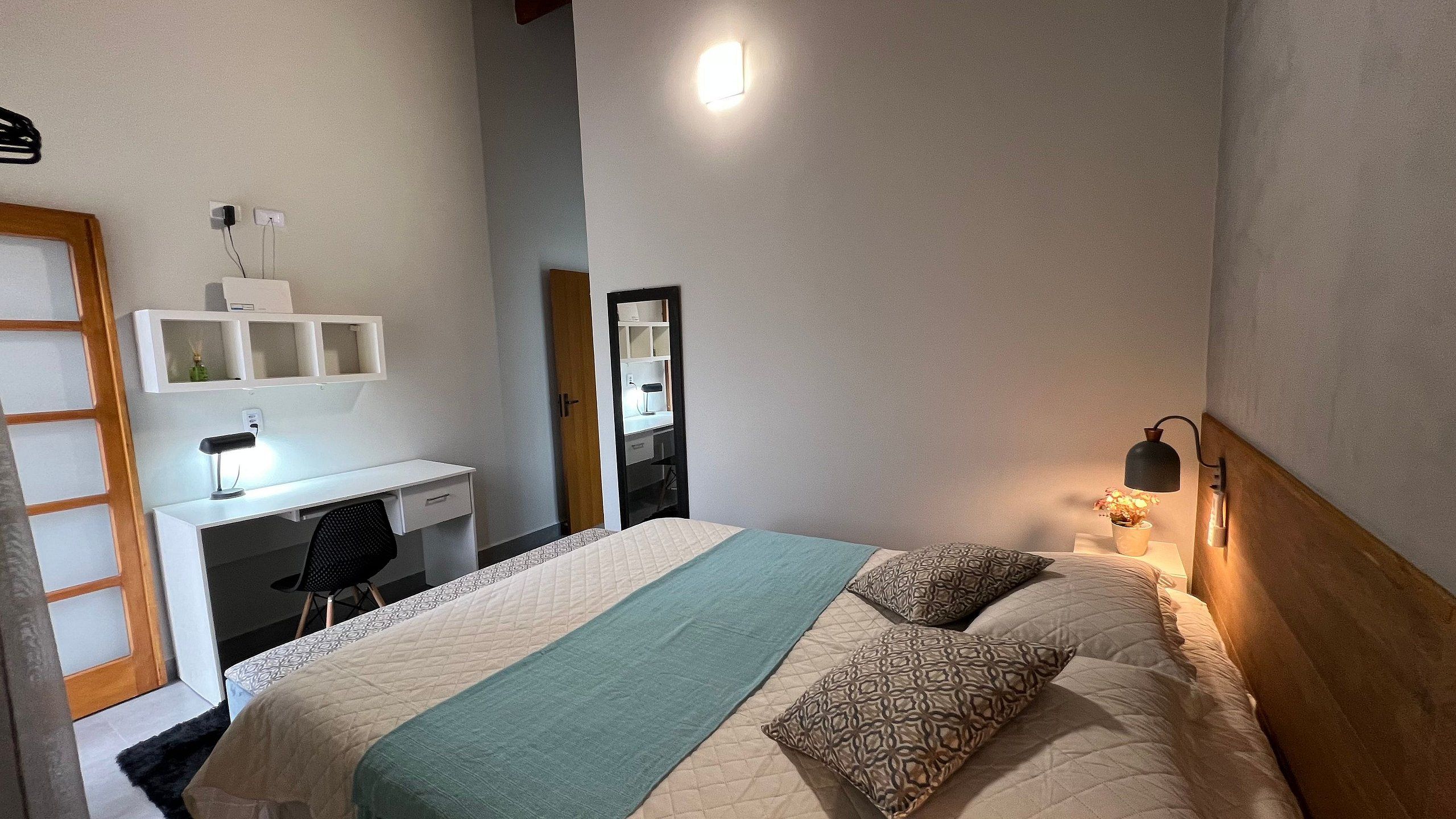 Un dormitorio con cama, escritorio, silla, espejo y mesita de noche con lámpara.