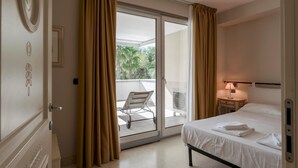 Une chambre d’hôtel avec un lit, une table de chevet, une lampe et une vue sur un balcon équipé d’une chaise et d’un ventilateur.