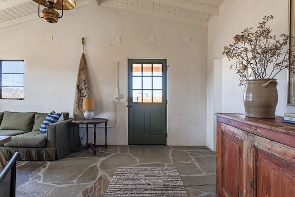 Une pièce avec un sol en pierre, une porte verte, un canapé avec des coussins, une lampe et un vase rempli de fleurs, posé sur une commode en bois.