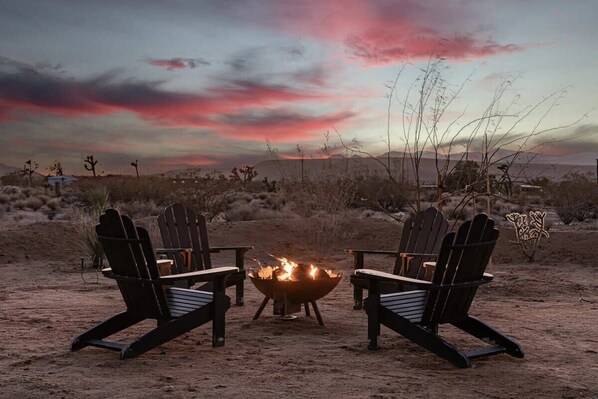 Twee Adirondack stoelen gericht op een kampvuur in een woestijnlandschap met een zonsondergang.
