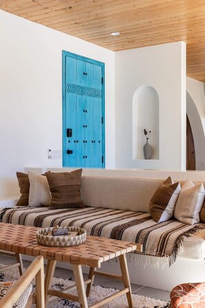 Una sala de estar acogedora con techo de madera, una puerta azul, un sofá color beige y una mesa de centro de fibras naturales.