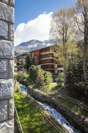 Un immeuble à plusieurs étages doté de balcons, entouré d’arbres et bordé par un ruisseau.