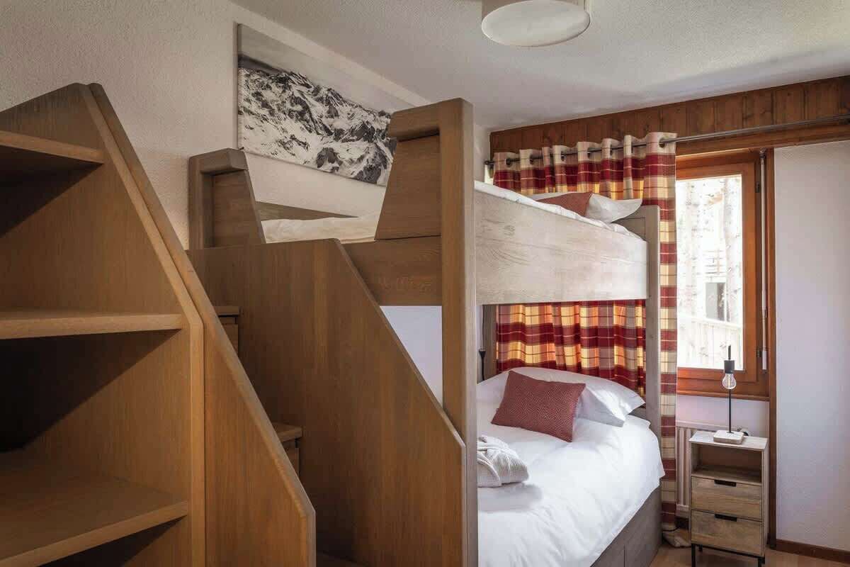 Uma cama de beliche com escada de madeira, uma mesa de cabeceira, um abajur e uma janela com cortinas.