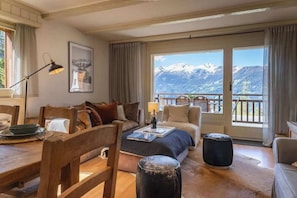 A cozy room with a large window, a sofa, a dining table, and a view of mountains. - 3BR Central: Pool, Sauna, Gym & Views (Verbier)
