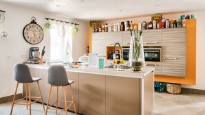 A modern kitchen with a central island, bar stools, a clock, and a variety of kitchen appliances and storage cabinets. - Stunning 4* Home for 6, Beach & Port Walkable (Le Guilvinec)