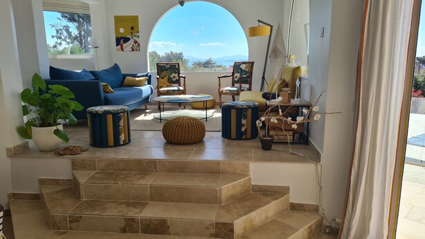 Un salon avec un canapé bleu, des poufs ronds et une grande fenêtre donnant sur la mer.