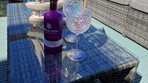 Pöydällä on gin-pullo ja lasi ulkona olevalla parvekkeella, jolta avautuu näkymä taloihin ja purjeveneeseen.