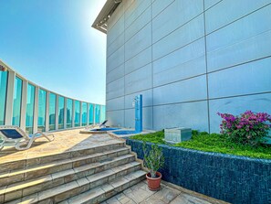 Un bâtiment moderne doté d’une grande façade en verre, d’un espace piscine avec des chaises longues et d’un escalier menant à l’entrée.