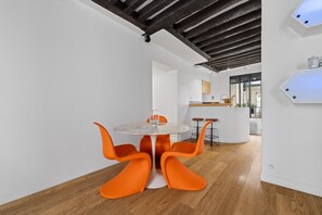 Uma sala de jantar moderna com uma mesa redonda e cadeiras laranjas, uma cozinha ao fundo e piso de madeira.