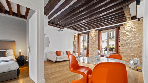 A modern room with a bed, white sofa, dining table with orange chairs, and a stone wall. - Lucius, 1BR/1BA, 2 people (París)