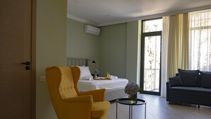 A hotel room with a bed, a yellow armchair, a gray sofa, a small table with a vase, and a window with curtains.