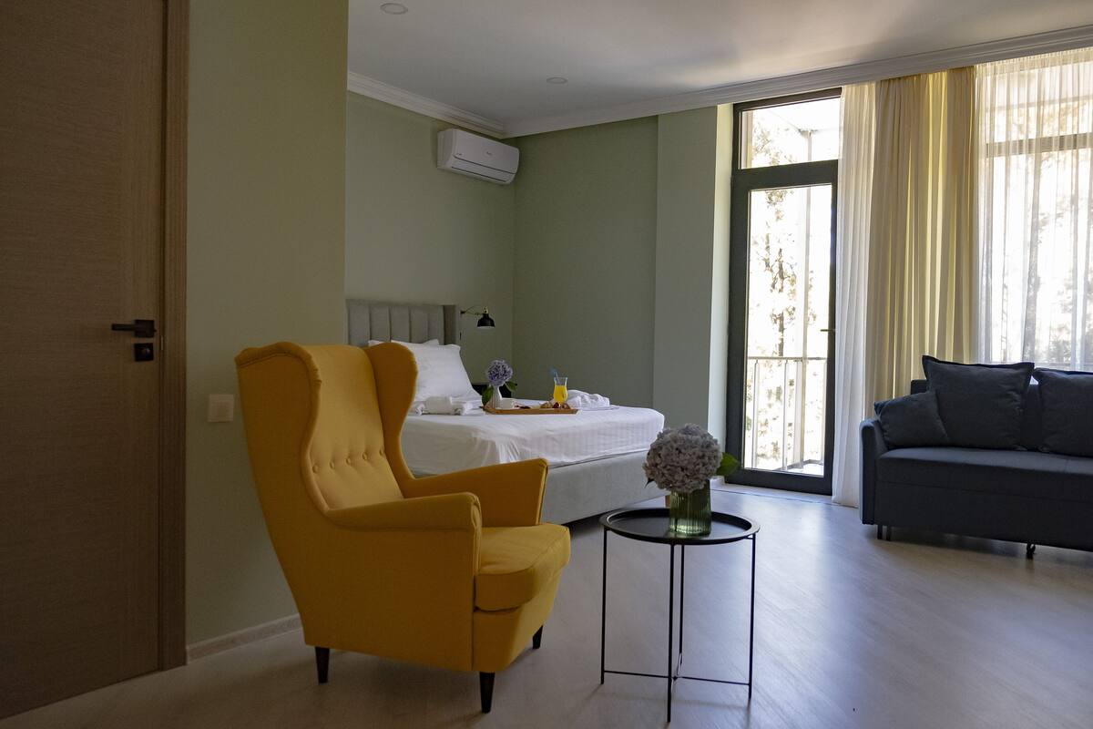 Une chambre d’hôtel avec un lit, un fauteuil jaune, un canapé gris, une petite table avec un vase et une fenêtre avec des rideaux.