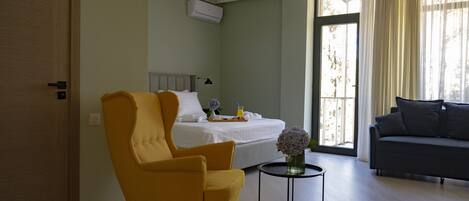 A hotel room with a bed, a yellow armchair, a gray sofa, a small table with a vase, and a window with curtains.