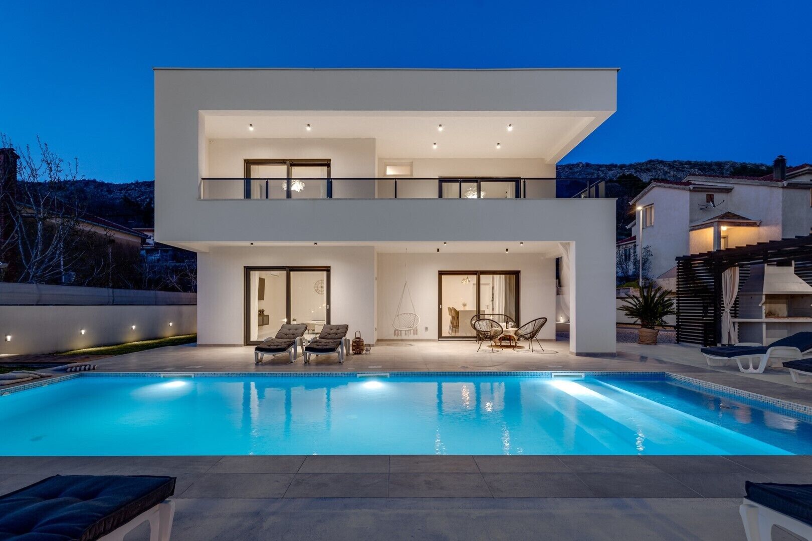 Ein modernes, zweistöckiges Haus mit Swimmingpool, Sitzgelegenheiten im Freien und einem Balkon.