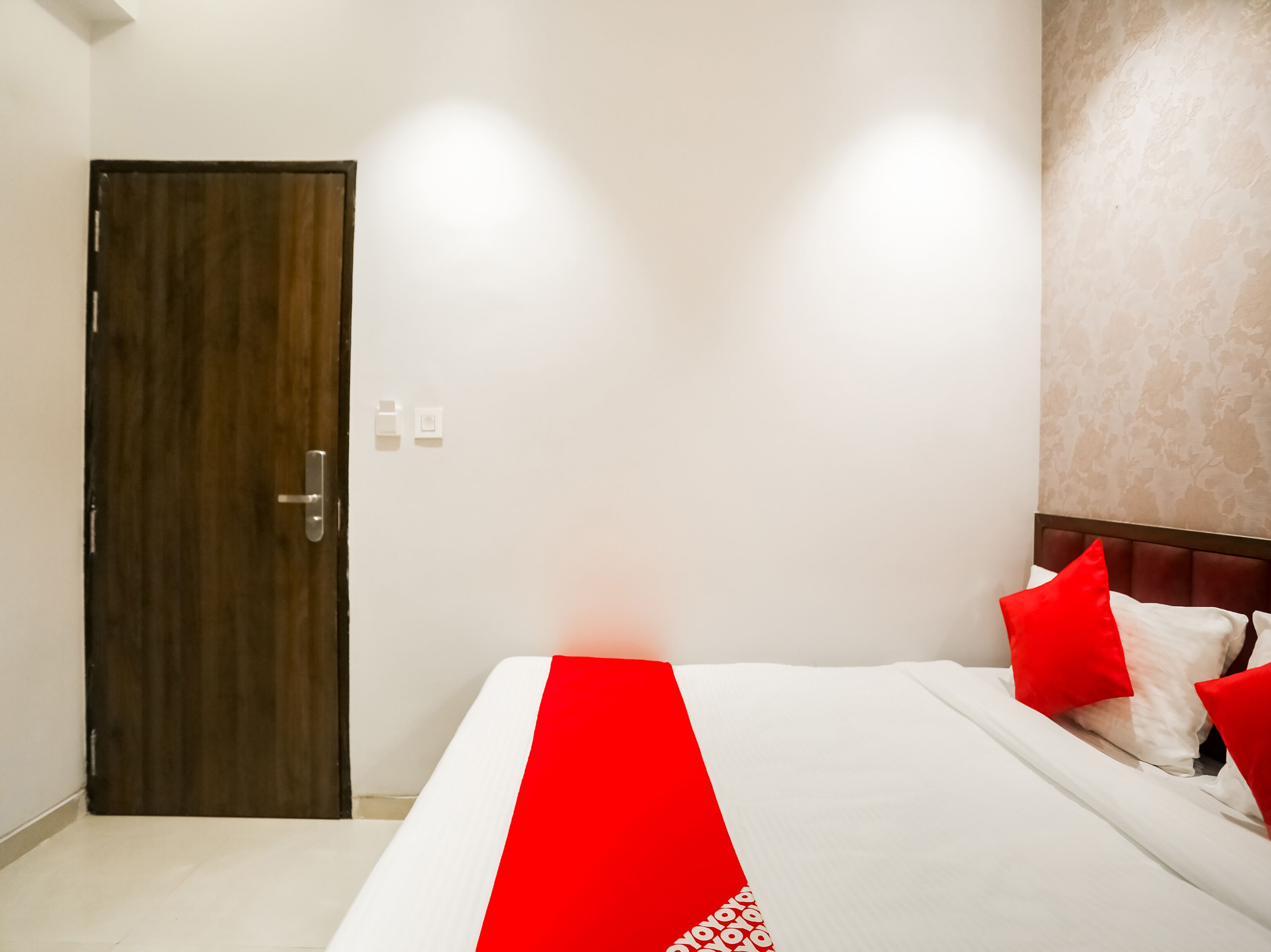 A hotel room with a wooden door, white walls, a bed with red and white bedding, and a patterned wall.