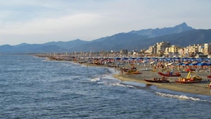 Una playa costera con numerosos sombrillas y barcos, un cielo despejado y montañas al fondo.