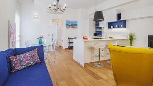 Un apartamento moderno con una sala de estar que incluye un sofá azul y un sillón amarillo, una cocina con armarios blancos y una lámpara colgante negra, y un comedor con una mesa de vidrio y sillas.