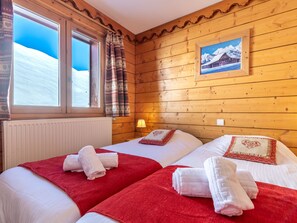 A cozy room with wooden walls, two beds with red and white bedding, a window with curtains, and a framed picture on the wall.