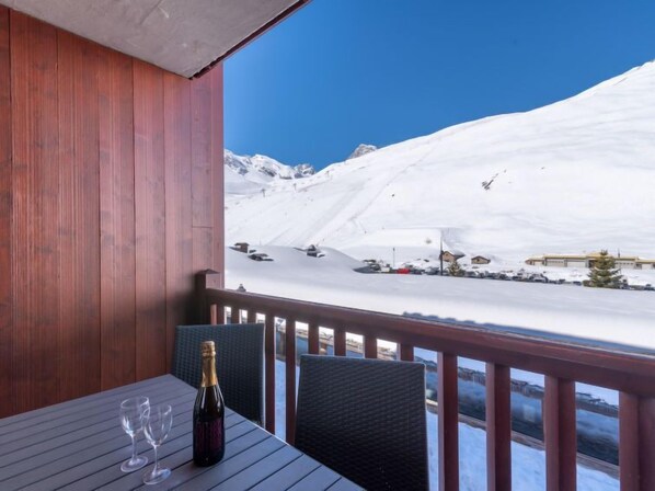 A wooden deck with a table and chairs, overlooking a snowy mountain with ski slopes and buildings.