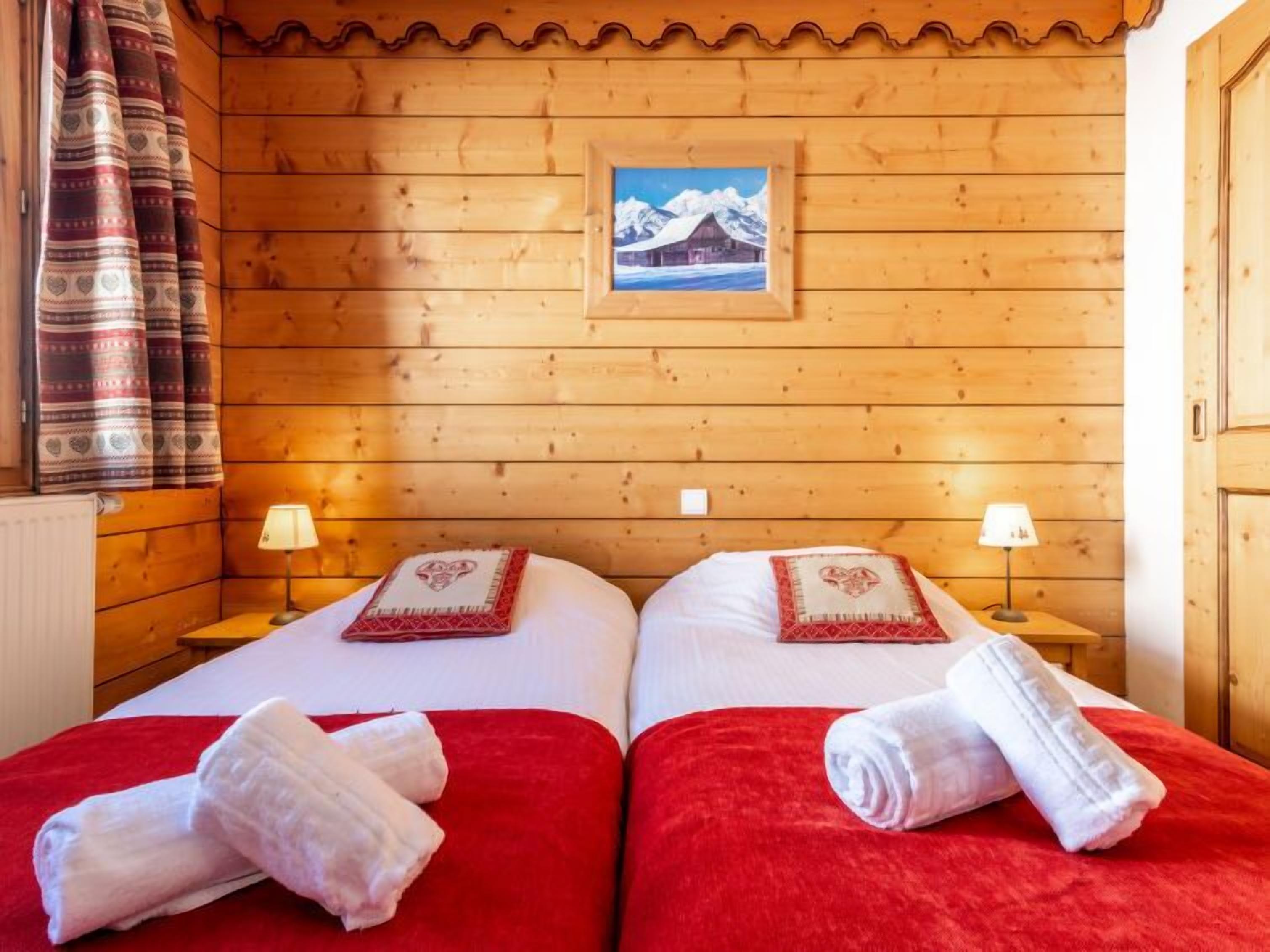 A wooden cabin room with two beds, red bedding, and a painting of mountains on the wall.