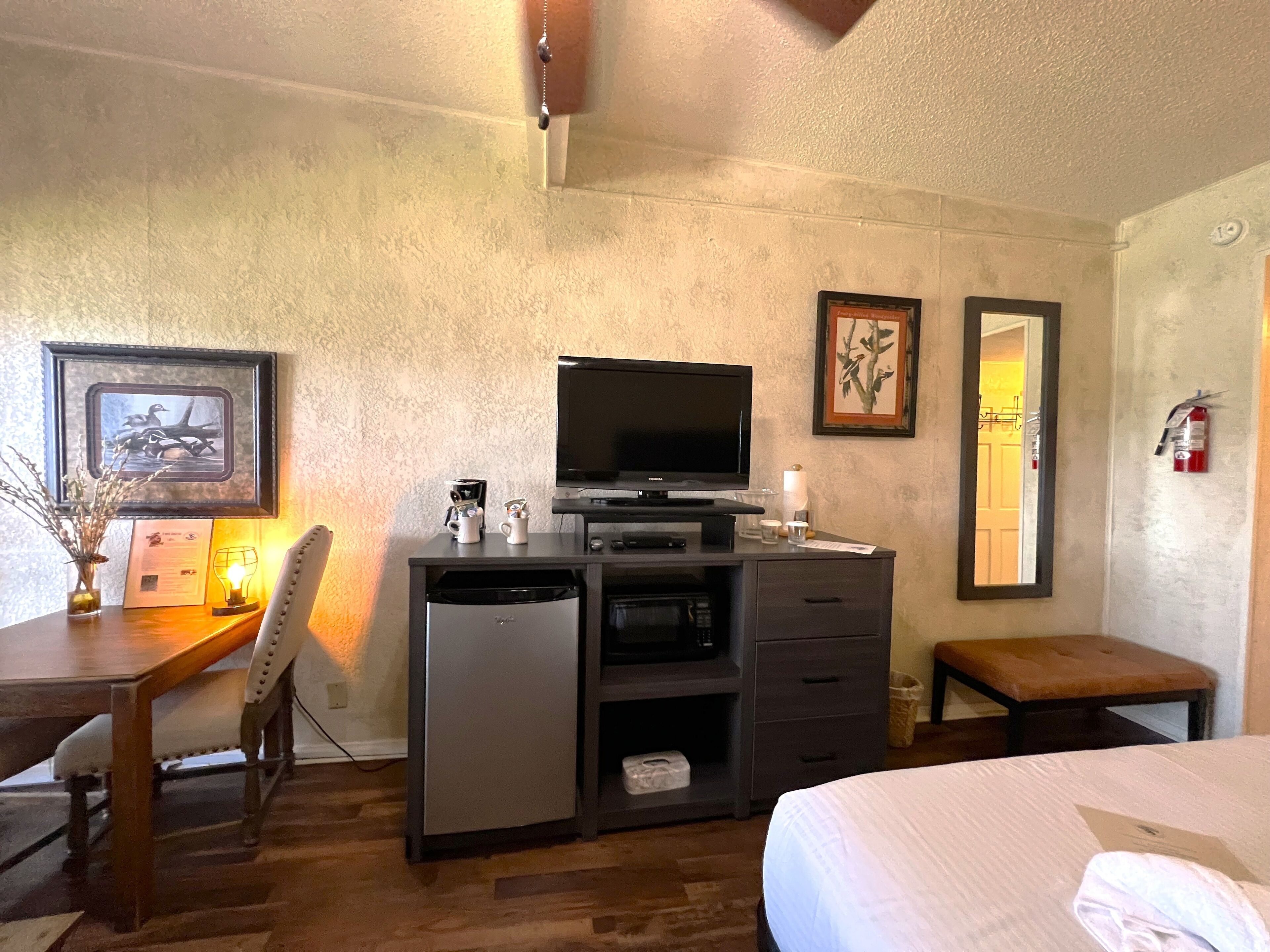 A compact hotel room with a bed, a desk with a television, a small seating area, and a mirror.