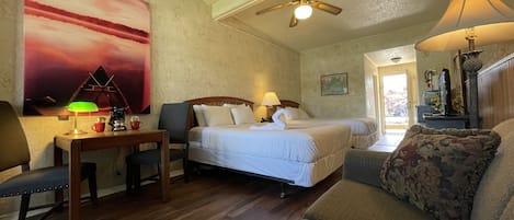 A hotel room with two beds, a desk with a lamp, a chair, a sofa, a ceiling fan, and a painting on the wall.