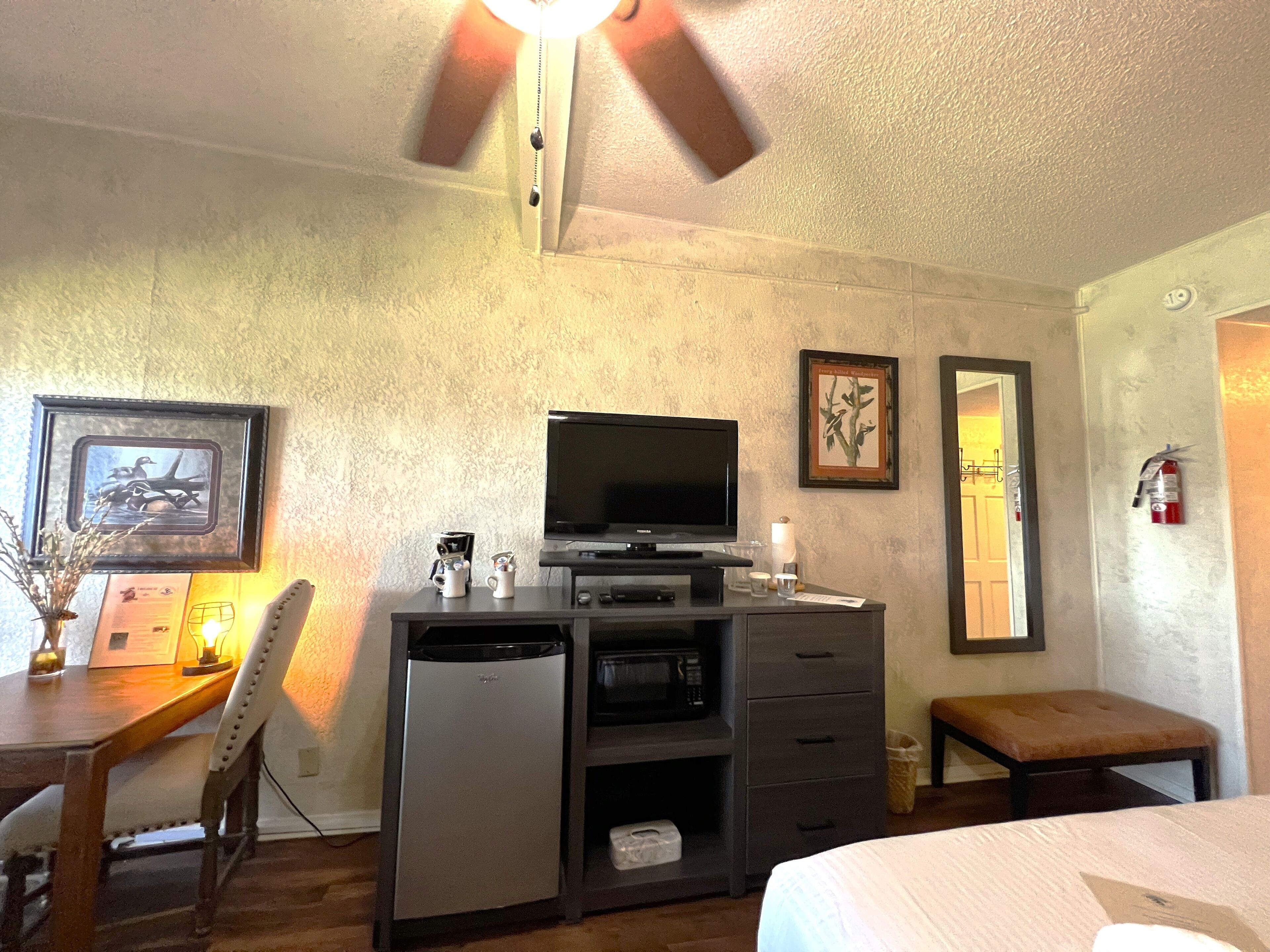 A compact hotel room with a bed, a desk with a television, a microwave, and a small refrigerator. There is a chair, a bench, and a framed picture on the wall.