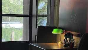A hotel room with a bed, a desk with a lamp, a chair, a window with a view of trees, and a painting on the wall. - Mountain Harbor King Guest Room on Lake Ouachita by RedAwning (Mount Ida)